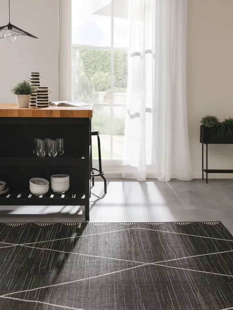 A spacious room with large windows draped in sheer curtains, a dark wooden sideboard with dishes underneath, and a geometric-patterned dark rug on a grey floor.
