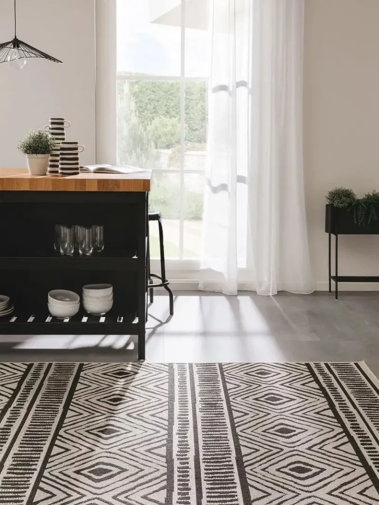 A well-lit dining area featuring sheer curtains, a wooden table with black legs, and a black and white geometric rug on a grey tiled floor.